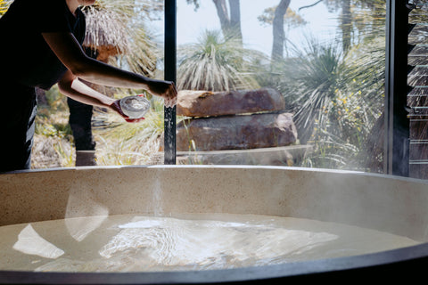 Person applying luxury bath salts to a large bath tub with a natural scene in the background.