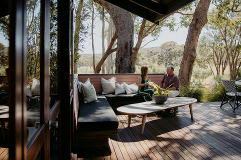 Two people sitting on a wooden deck with outdoor furniture and trees in the background