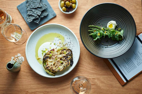 Two plates of gourmet food on a wooden table with glasses and a menu.