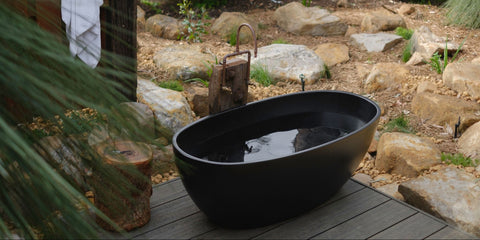 Black freestanding bathtub in a natural outdoor setting with rocks and wooden planks.