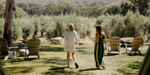 Two women walking on a grassy area with outdoor furniture and olive trees in the background