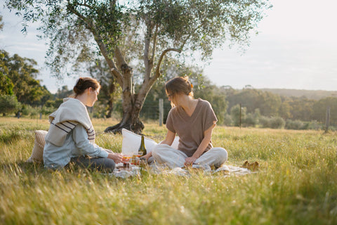 Two people sitting on a blanket in a grassy field with trees in the background