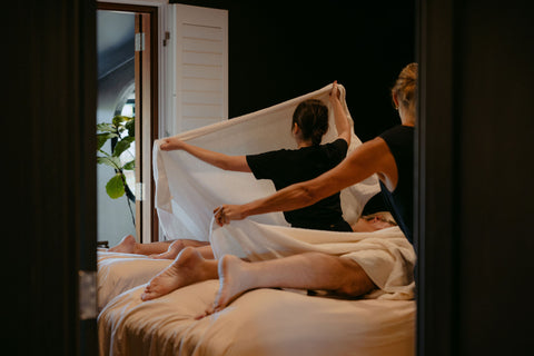 Two people lying on a bed with one person holding a white sheet over them.