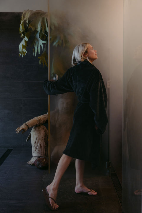Person in a dark robe standing in a dimly lit room with a plant and sculpture.