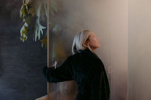 Woman in a bathrobe entering a spa steam room with a plant and a textured wall.