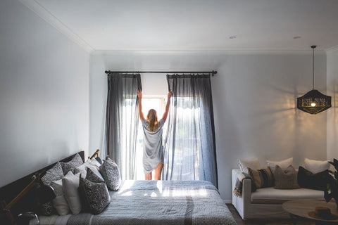 Woman opening curtains in a modern bedroom with a bed and sofa.