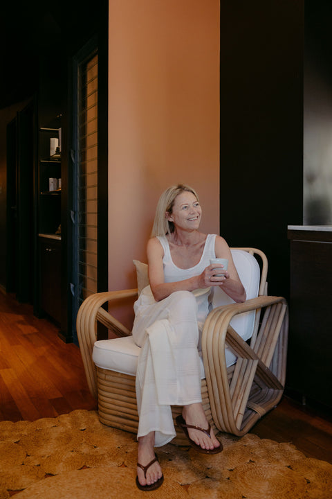 Woman sitting in a wicker chair holding a cup in a warm-toned room.
