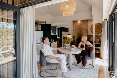 Two people sitting at a table in a modern living room with large windows.