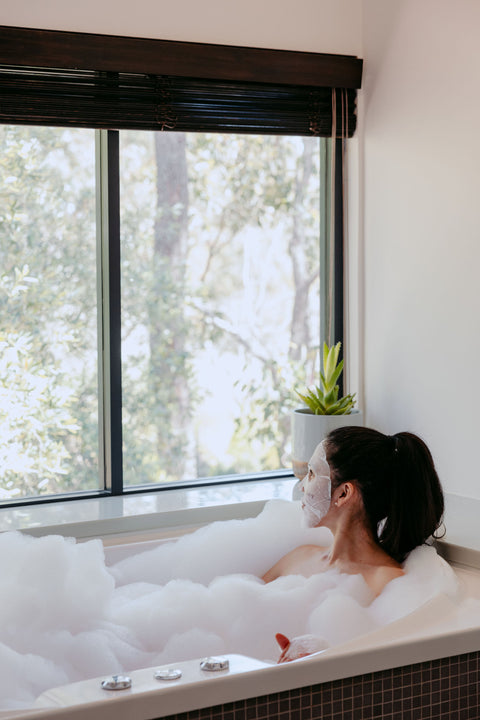 Woman relaxing in a bubble bath with a large window showing trees outside.