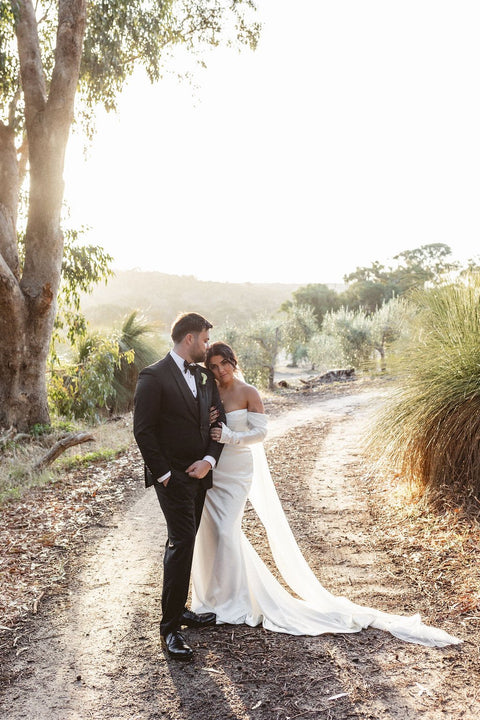 Couple in formal attire standing on a dirt path with trees and open space in the background