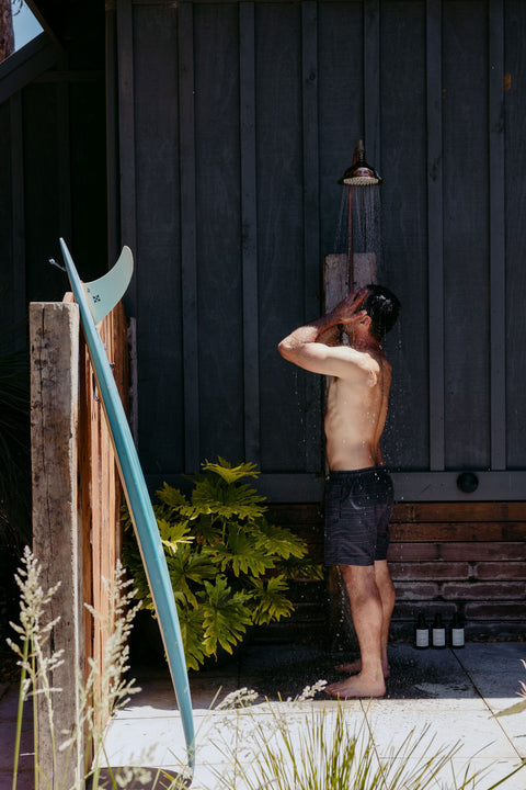 Person standing outdoors with a surfboard, under a showerhead.